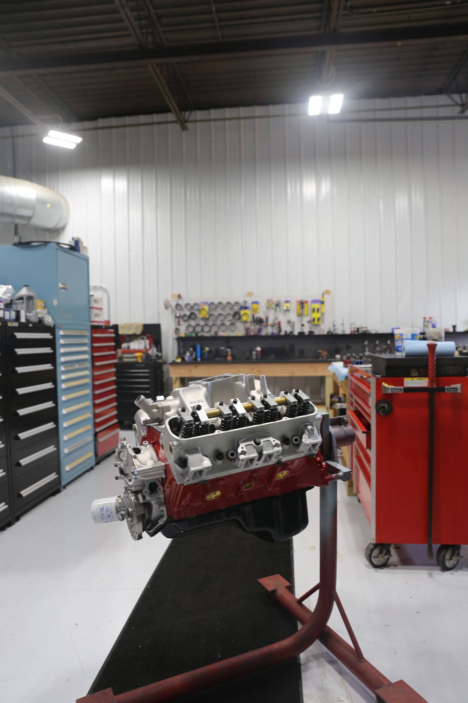 Engine block on a stand in a workshop, red and silver with tools and cabinets in background.