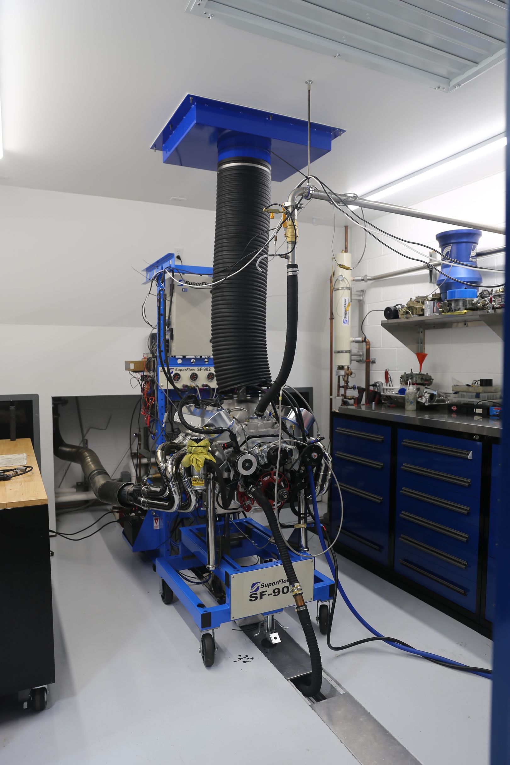 Engine test stand in a workshop, blue and black, with connected hoses and a large air intake.
