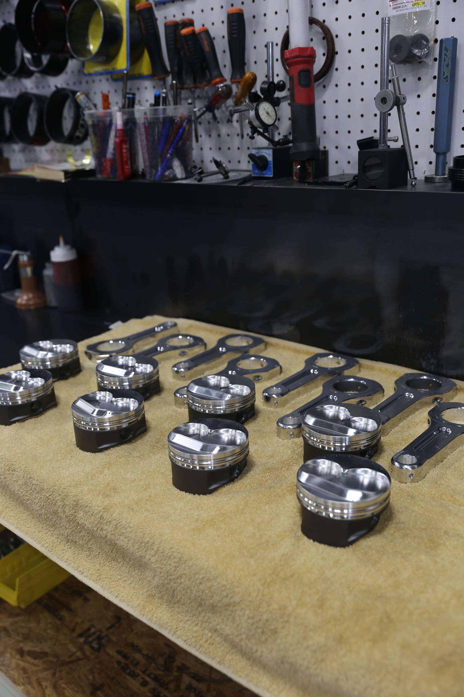 Twelve shiny metallic parts with connecting rods on a tan surface in a workshop setting.