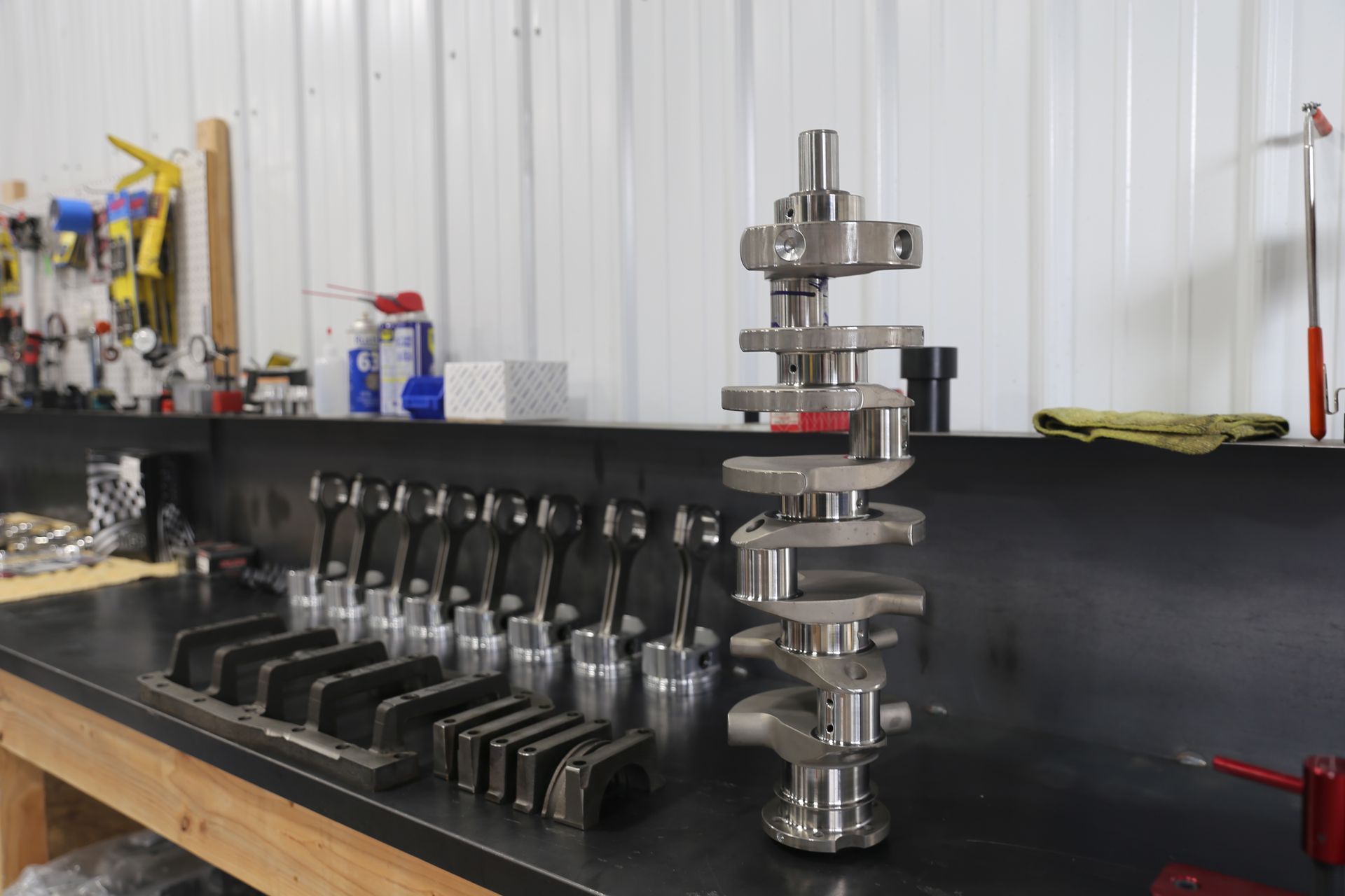 Crankshaft and engine parts on a workbench in a workshop. Parts are metal and gray; workbench is black.