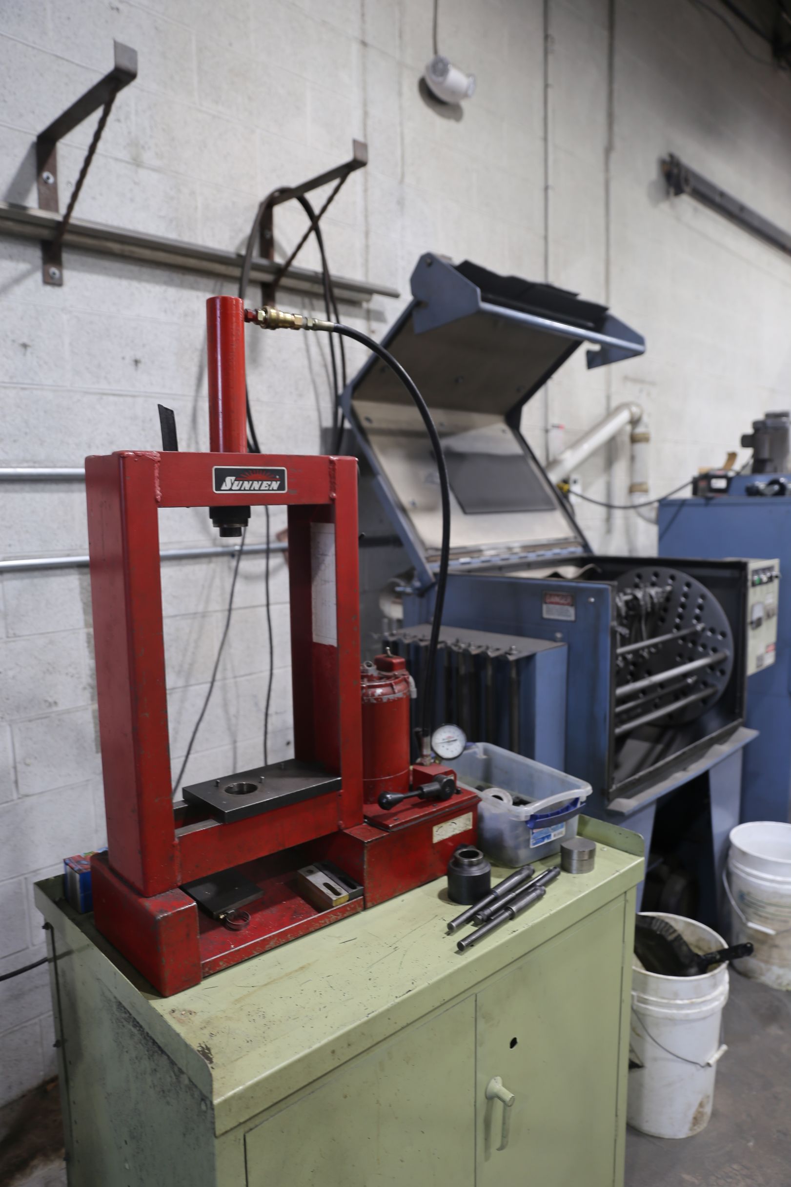 Red hydraulic press on a green cabinet in a workshop, with other industrial equipment nearby.