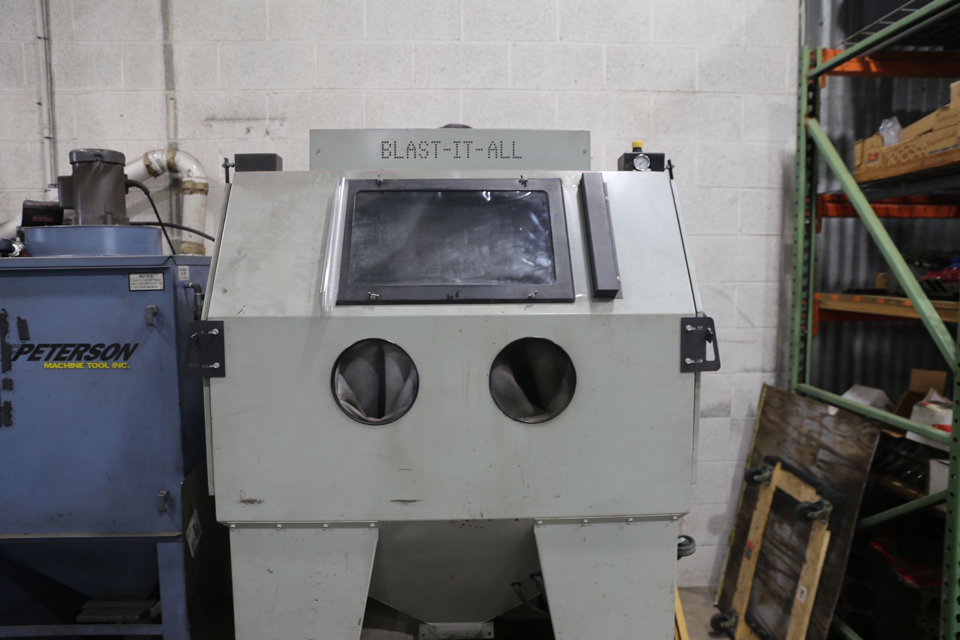 Sandblasting cabinet in a workshop, grey with a window and armholes. A blue dust collector is to the left.