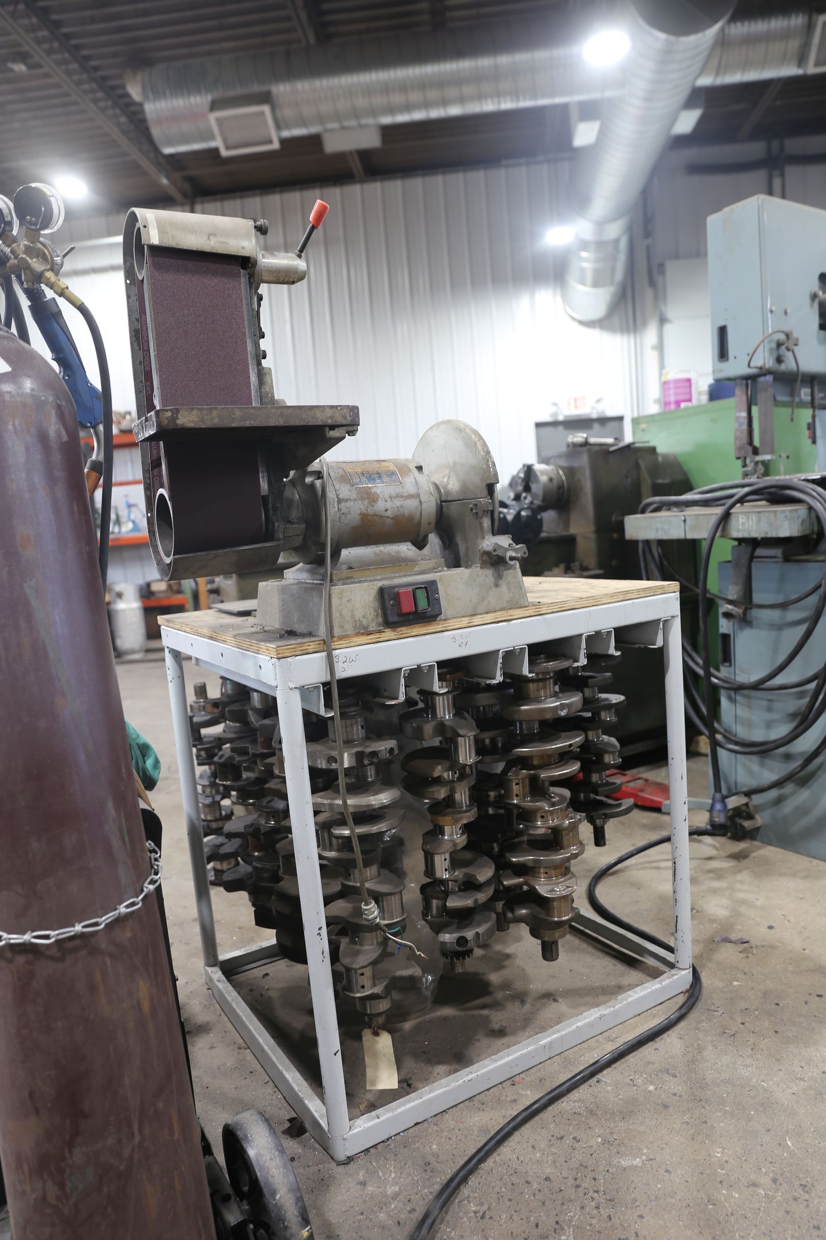A belt sander mounted on a metal cart filled with engine crankshafts in a workshop.