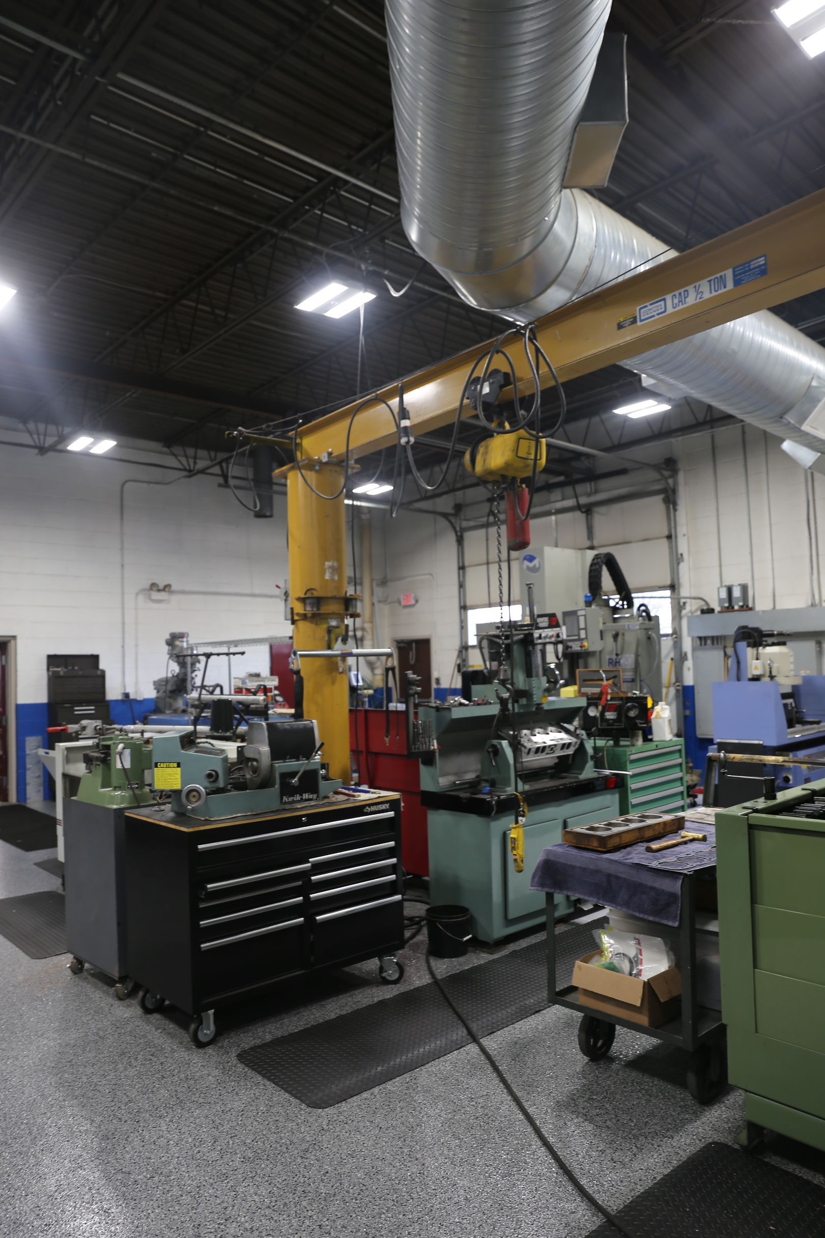 Workshop interior with machinery, tools, and a yellow overhead crane system. Grey floor and dark ceiling.
