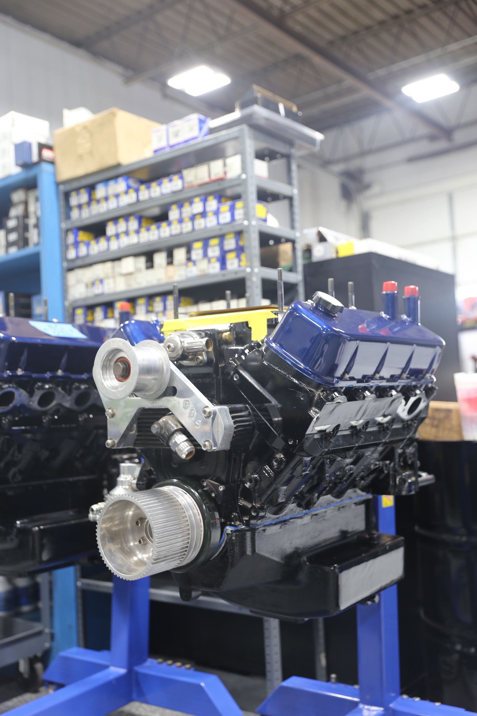 Dark-blue engine, silver bracket, and cylinder heads on a blue stand in a workshop with shelves of parts.