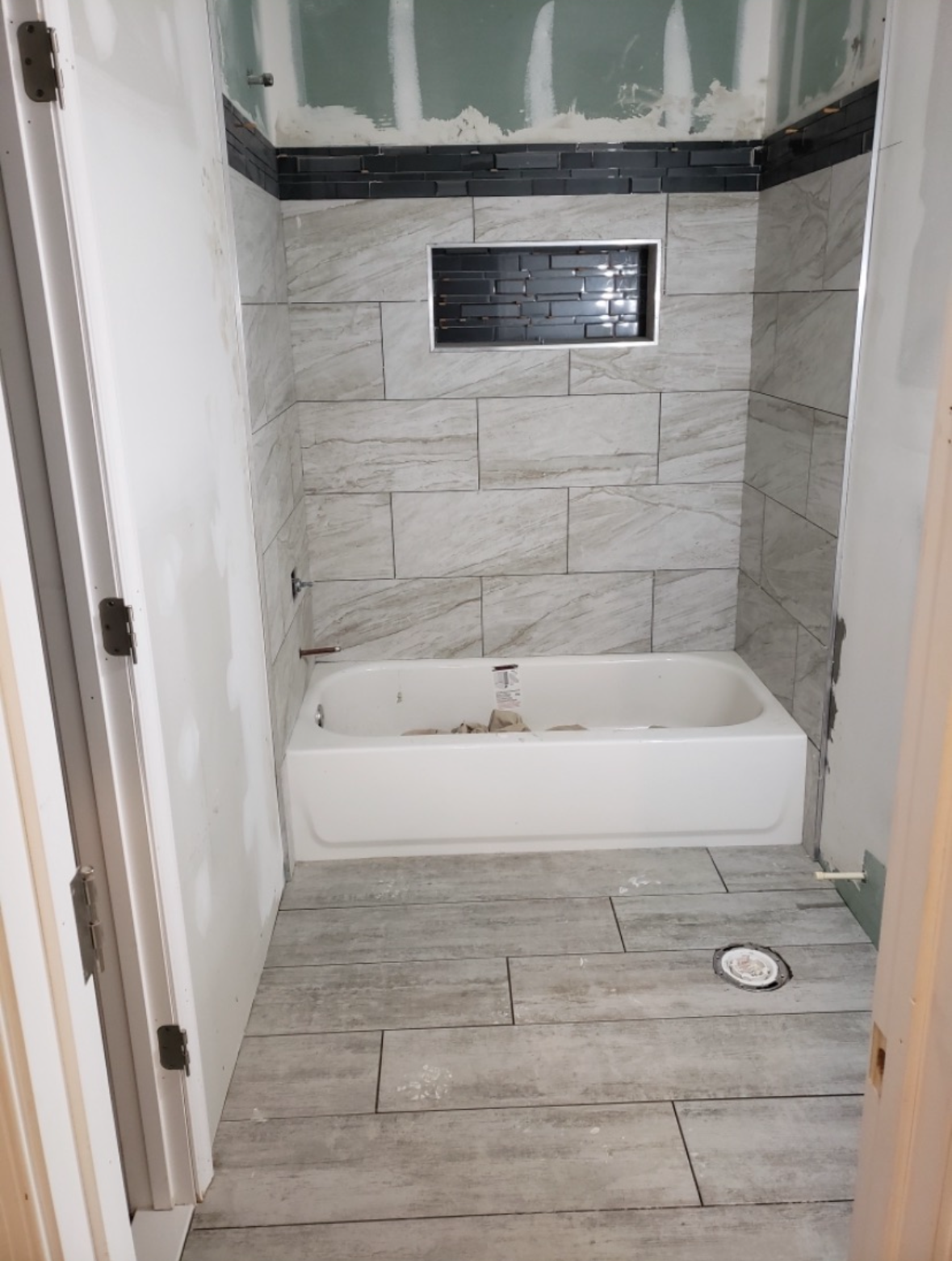 Bathroom under construction, tiled walls with a tub and inset shelf, grey and white tones.