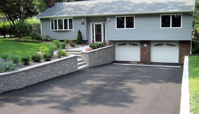 Gray house with a black asphalt driveway, a brick garage, and a stone retaining wall.