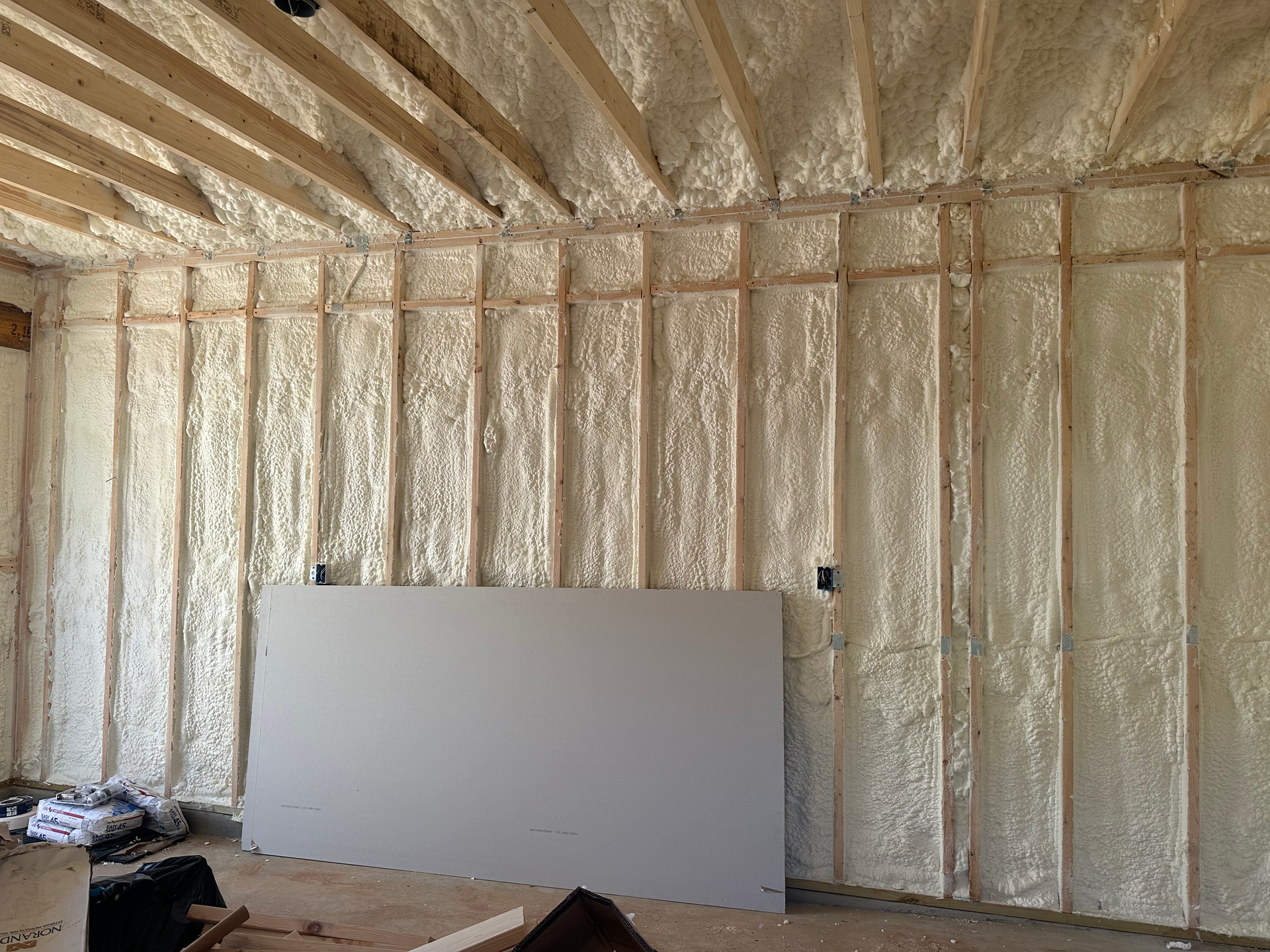 Interior wall construction with spray foam insulation. Beige foam fills wooden framing. Drywall partially installed.