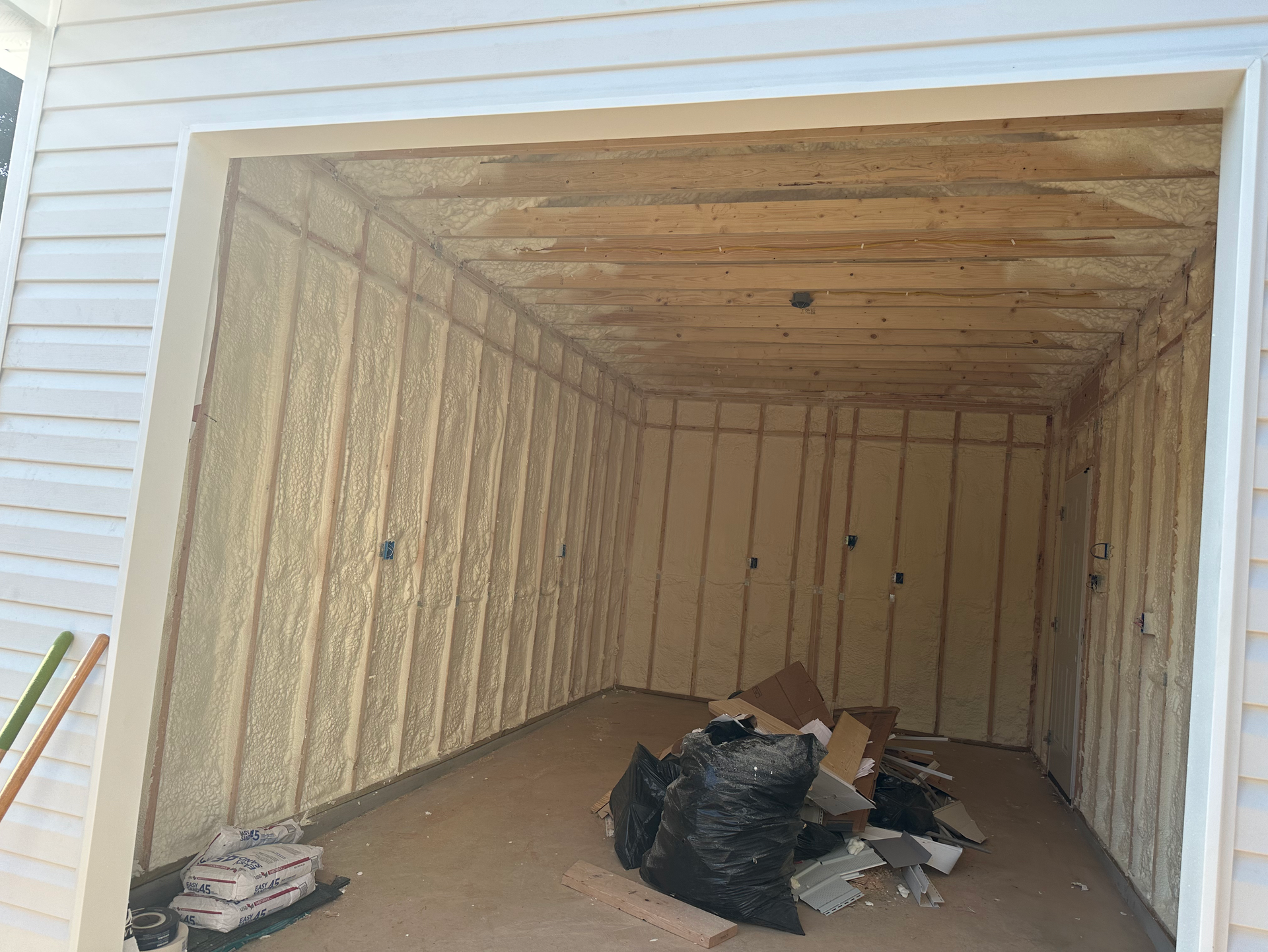 Garage interior with tan foam insulation, a black trash bag, and debris on the floor.