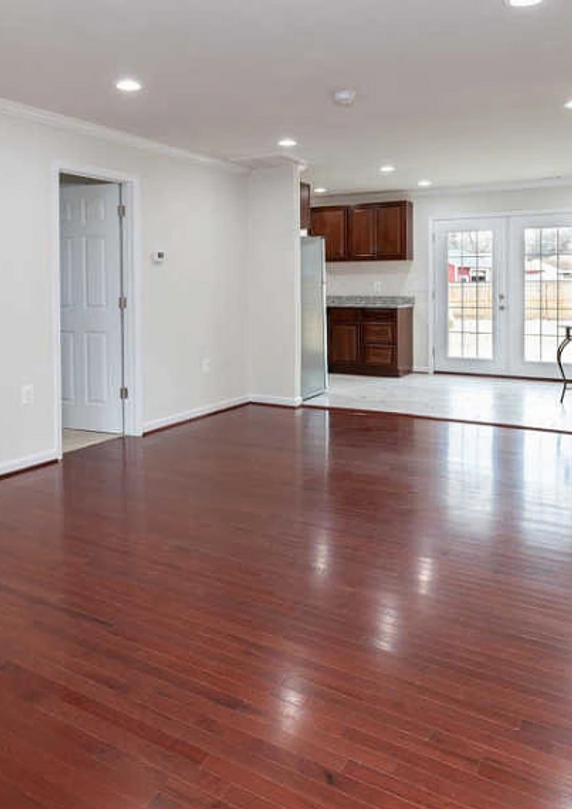Spacious living area with dark wood floors, white walls, and a glimpse of the kitchen and French doors.