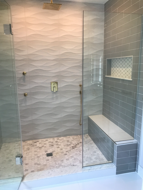 Modern shower with gray wave-patterned wall tiles, glass doors, gold fixtures, and a bench.