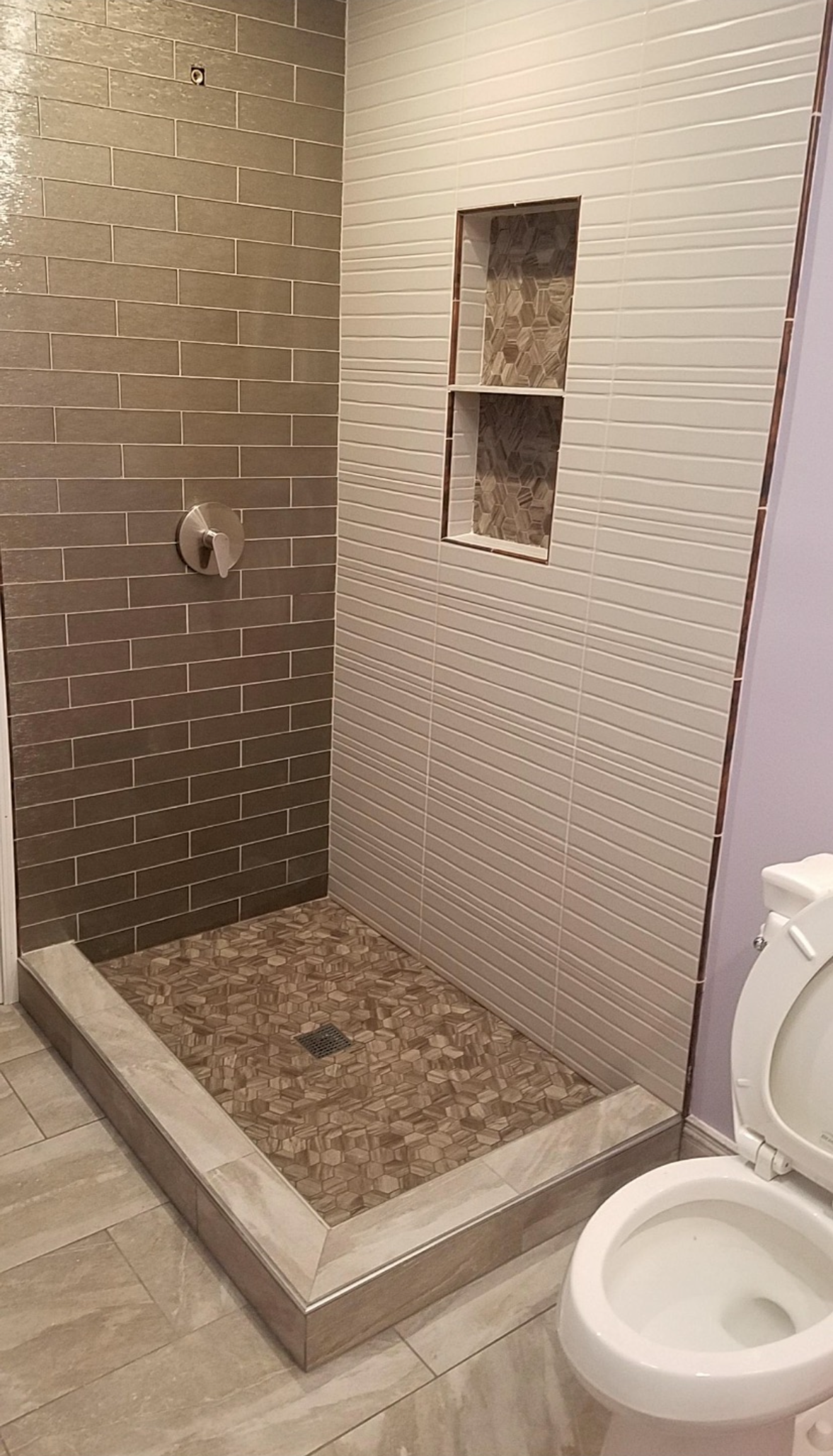 Shower stall with textured, beige tile and built-in shelf. Toilet in the foreground, light purple wall.