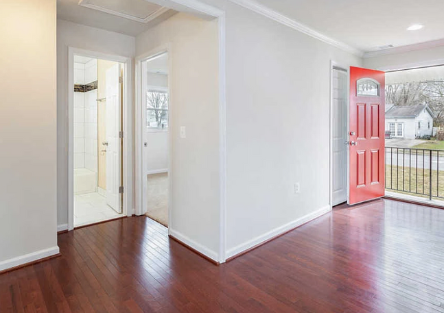 Interior view of a home with wood floors, an open red door, and a hallway leading to a bathroom.