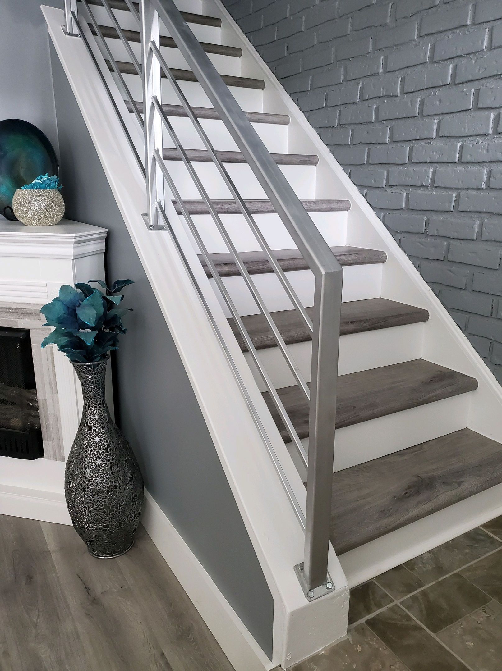 Staircase with gray treads, white risers, and silver railing, with a gray brick wall in the background.