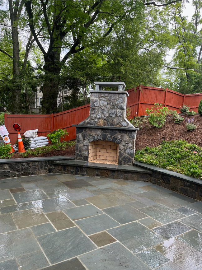 Stone fireplace on a patio with flagstone, orange fence and green foliage.