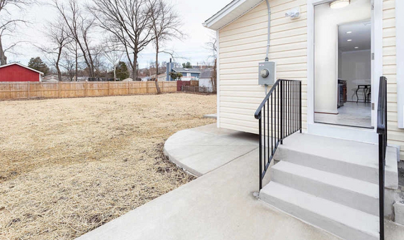 Back door with steps and railing, concrete path and yard. Fenced in with a red building in the distance.