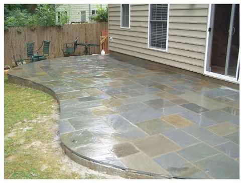 Stone patio extending from a house, bordered by grass and a wooden fence.