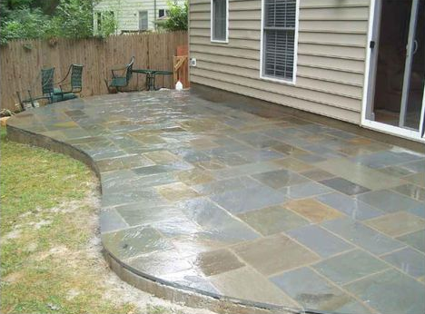 Stone patio next to a house. Curved edge borders the grass, two chairs, and a small table.