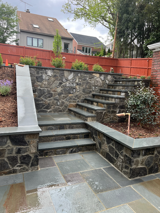 Stone steps and walkway in a landscaped yard, with a retaining wall and red fence in the background.
