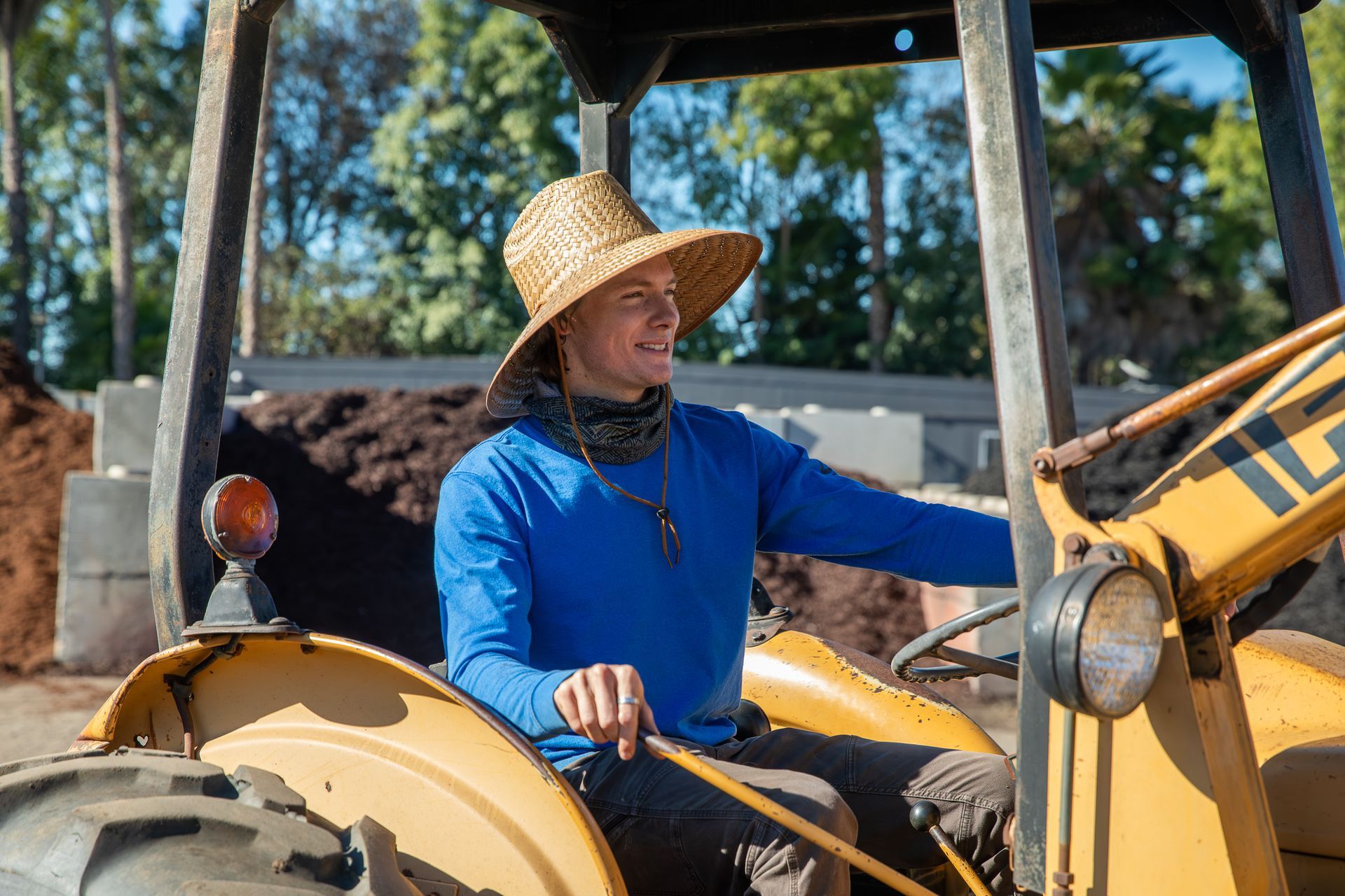 Person driving a yellow tractor, wearing a straw hat and blue shirt in an outdoor setting.