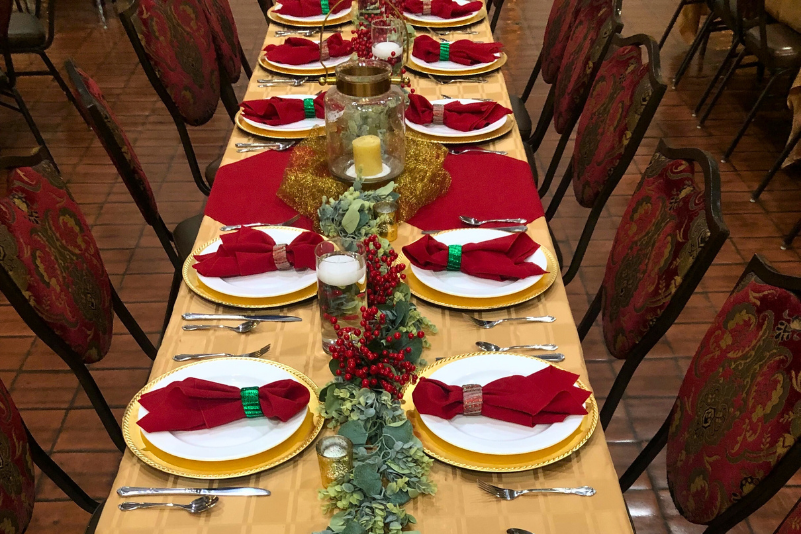 Banquet hall set for a holiday event with round tables covered in red cloths and festive greenery.