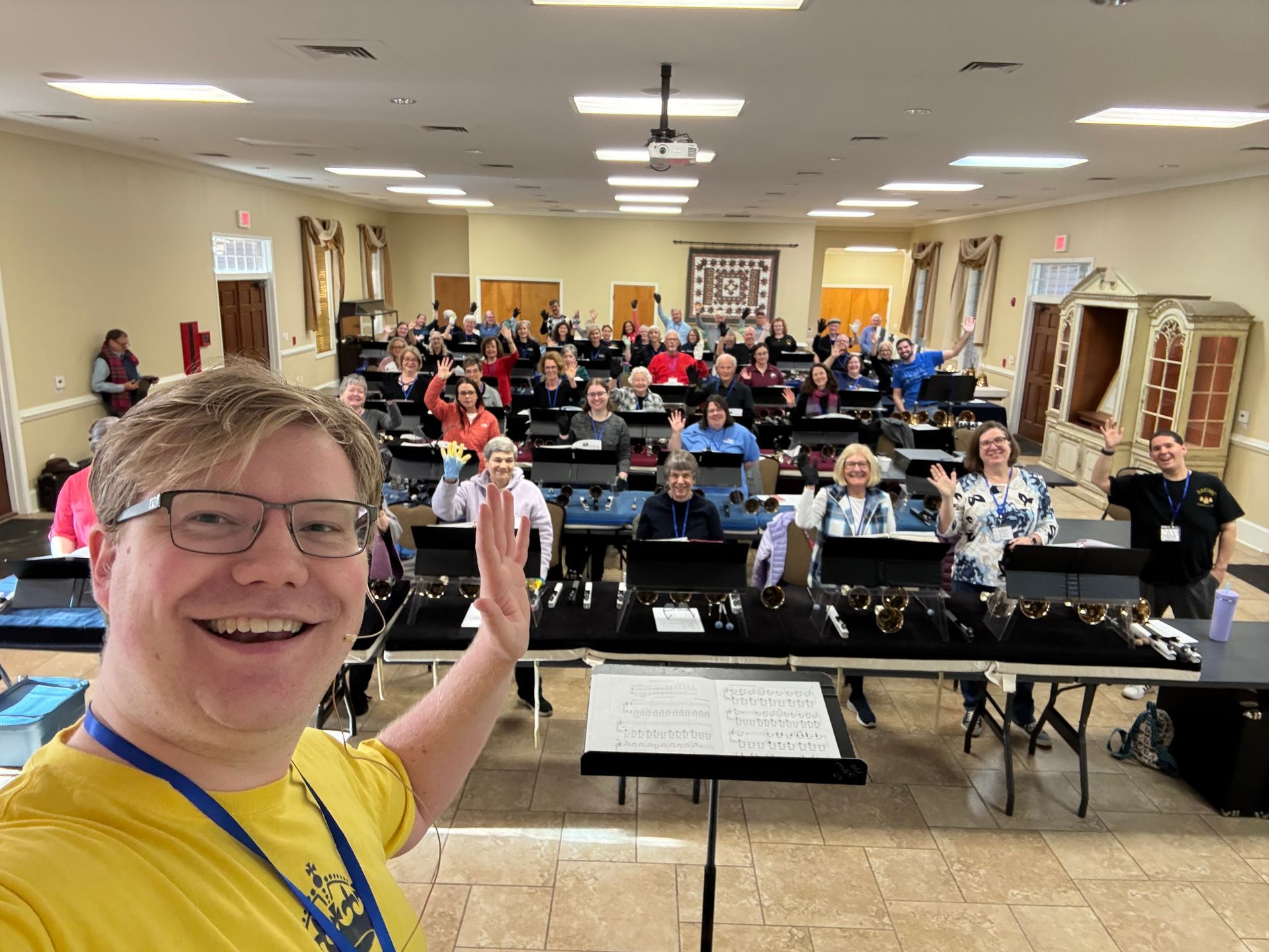 A handbell choir of 30+ people in a large room, ready to perform with a man wearing yellow shirt.
