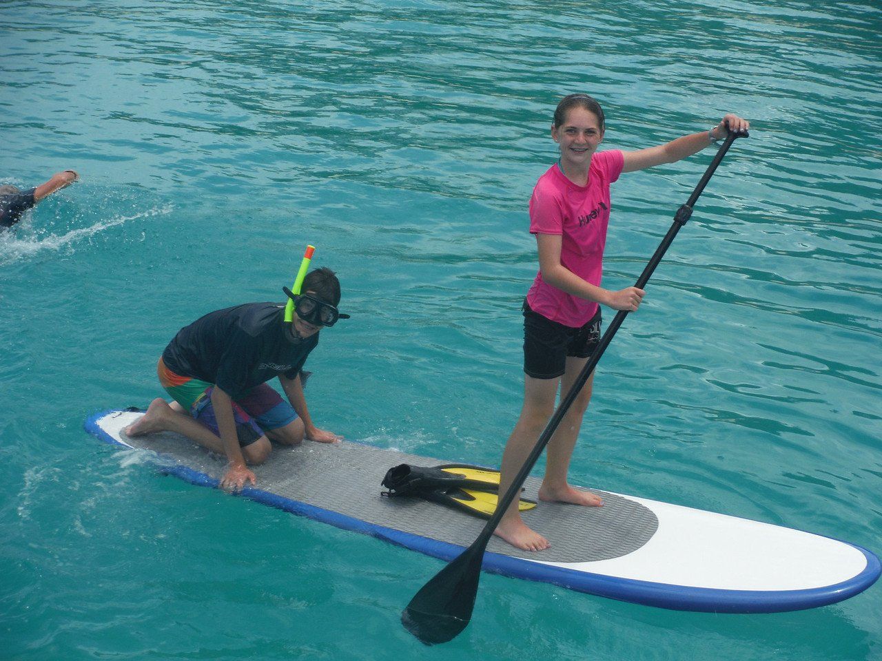 two people on a paddleboard, one sitting, wearing snorkel gear, another standing with the paddle