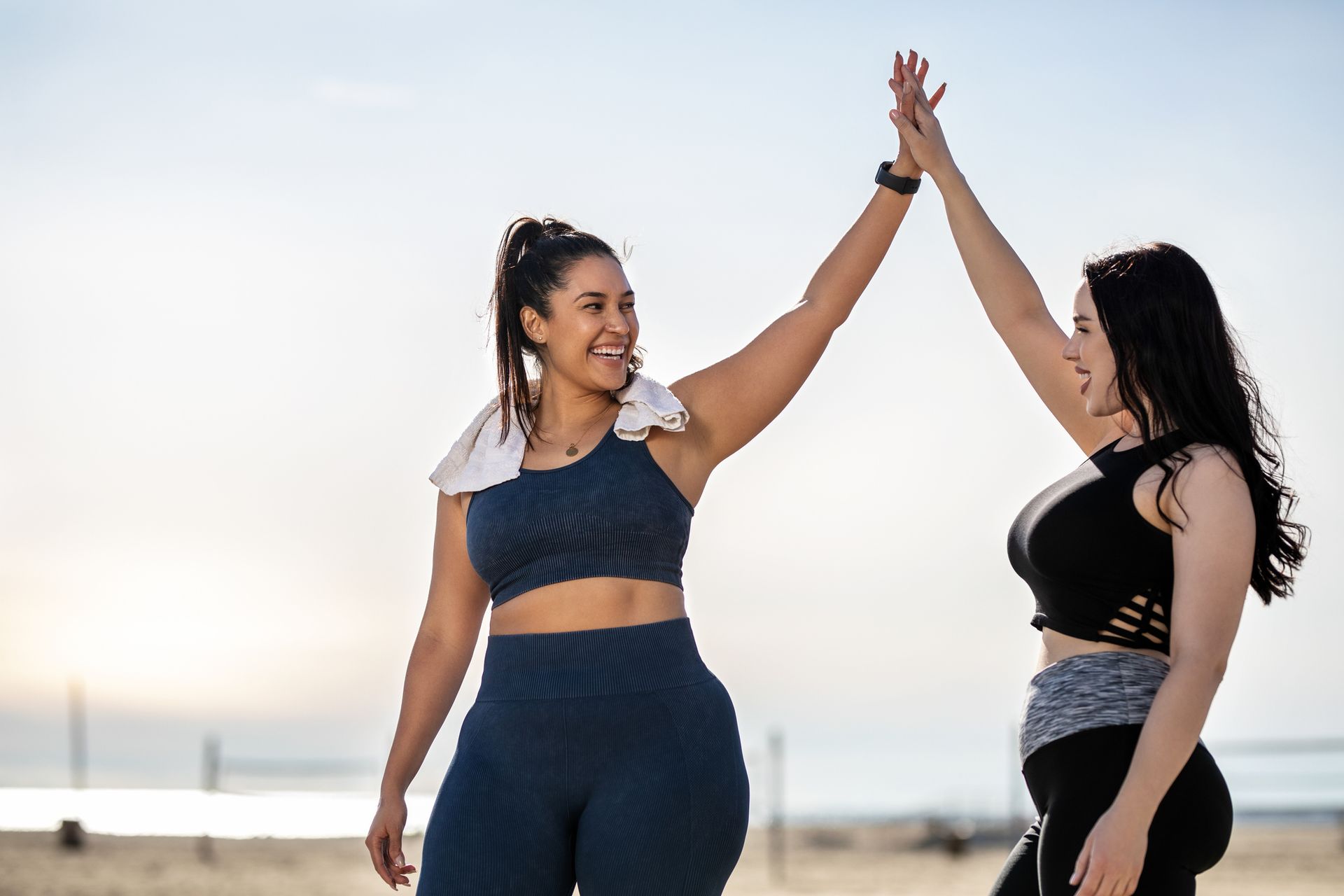 Two women in workout clothes high-fiving on a beach, smiling.