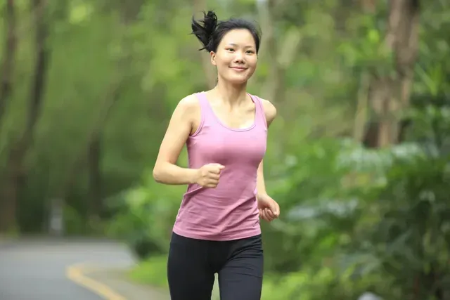 Woman running on a road through a green, tree-lined area, smiling. Wearing a pink tank top and black leggings.
