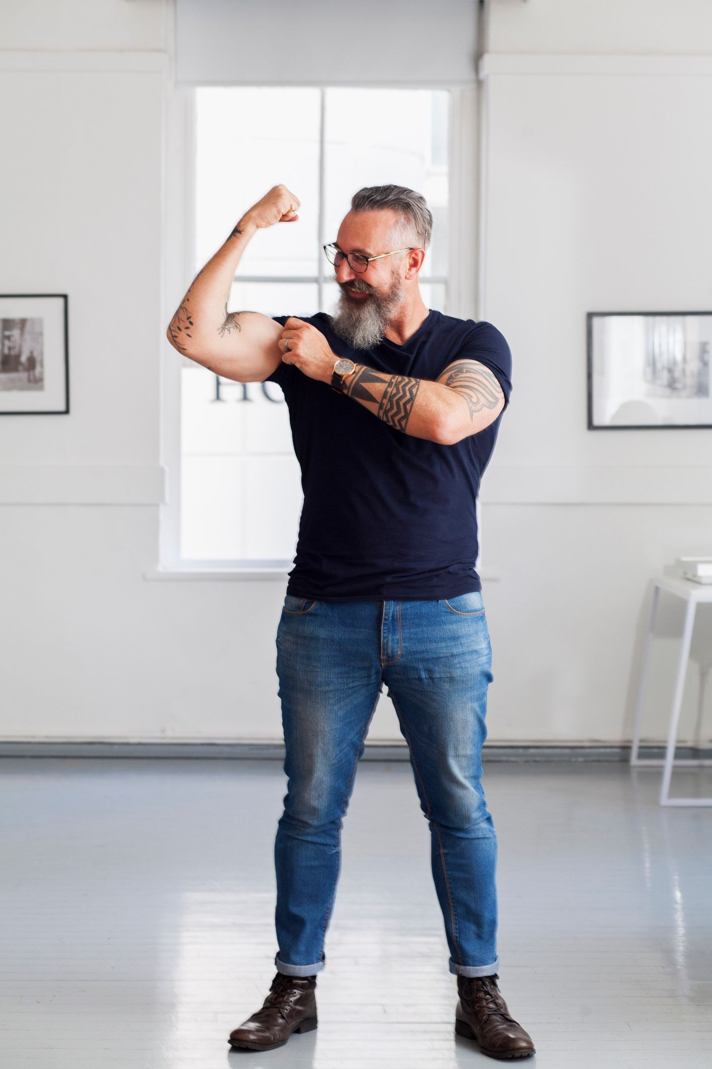 Man flexing arm, showing tattoos, smiling. Wearing jeans, dark shirt, and boots indoors.