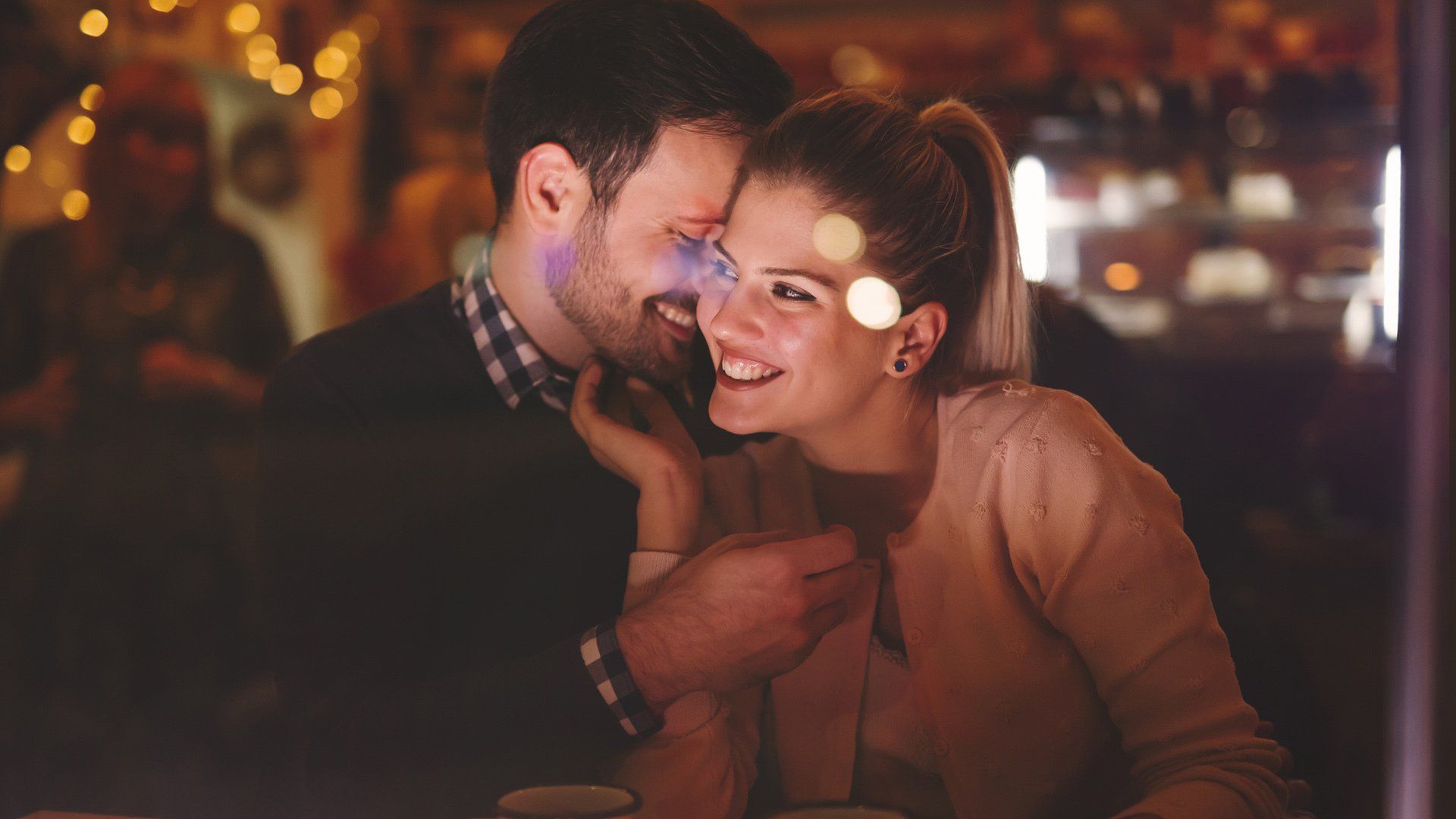 Couple in a dimly lit setting; woman smiles as man whispers into her ear, both appear happy.