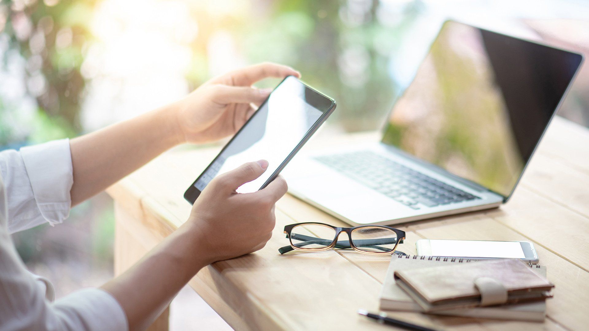 Person holding tablet, laptop open nearby on wooden table with glasses, notebook, and pen.