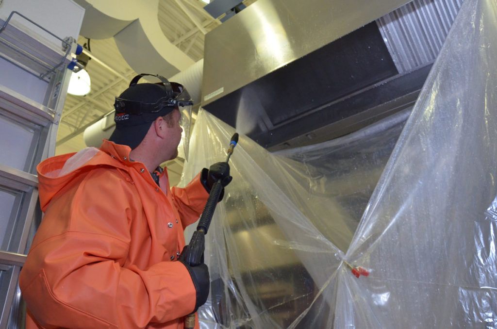 Man in orange protective gear power washing a stainless steel kitchen vent.