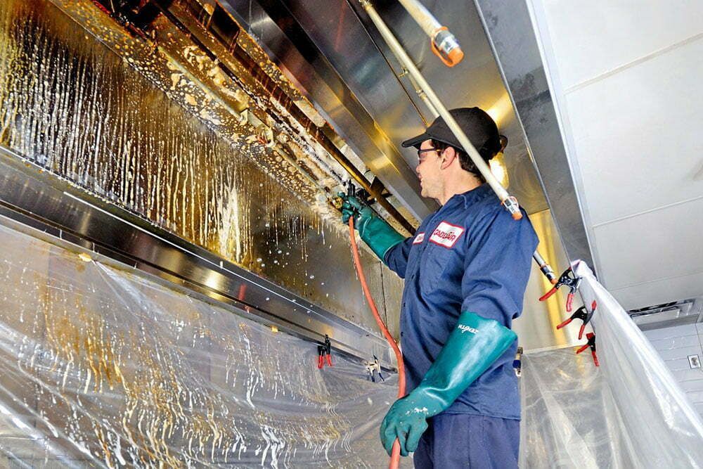 Man in work clothes cleans a greasy commercial kitchen vent hood with a spray nozzle.