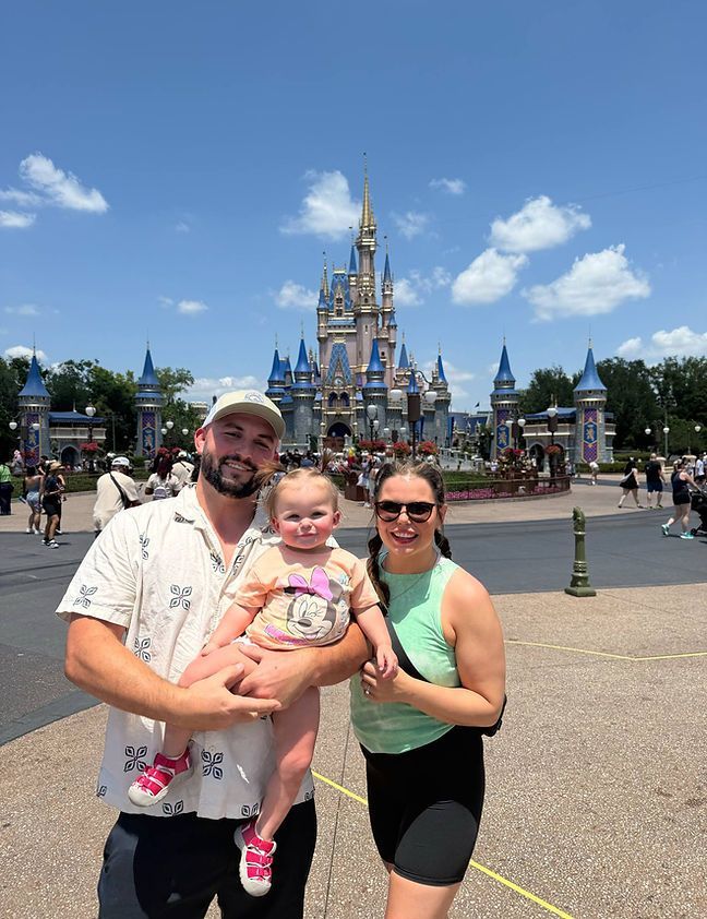 A family of three poses in front of Cinderella's castle at Disney World. The parents hold their young child, all smiling on a sunny day.