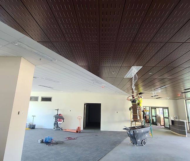 Interior under construction: Workers install a dark brown ceiling in a room with light-colored walls and concrete flooring.