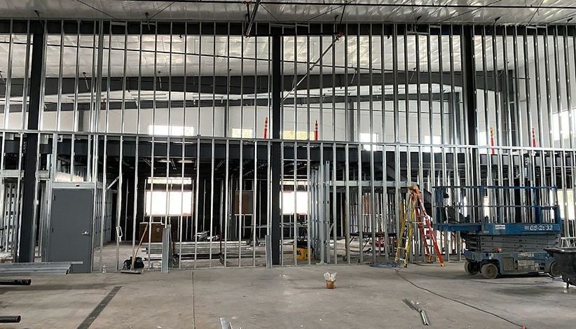 Interior view of a commercial space under construction, steel framing defines walls and openings. Construction tools and equipment are scattered around.