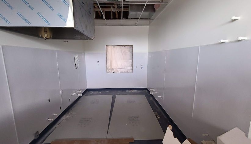 A kitchen under construction with white wall panels, a metal vent hood, and an unfinished floor. An empty window is visible at the center.
