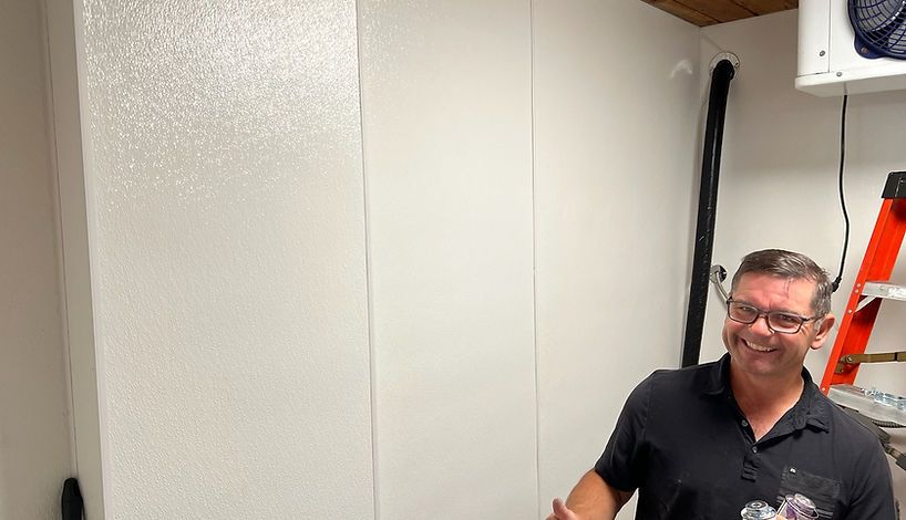 A man smiles in a white-walled room. He wears glasses and a black shirt, giving a thumbs-up.