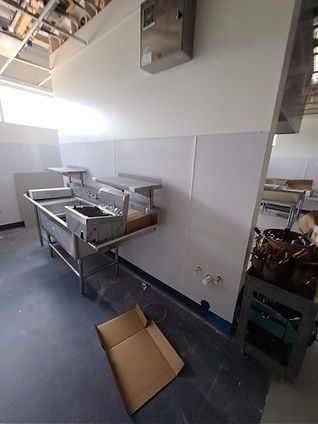 A commercial kitchen under construction, featuring a stainless steel sink and countertop against a white wall.