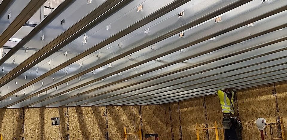 A person in a high-visibility vest works on a metal ceiling frame. The construction site has exposed wood walls.