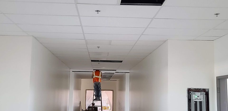A worker on a ladder repairs a suspended ceiling in a white hallway. The worker wears an orange safety vest.