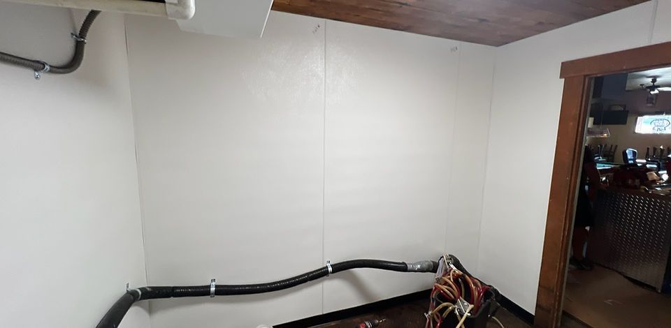 A small, white-walled room with a dark doorway, black pipes, and exposed wires, likely a utility or storage space.