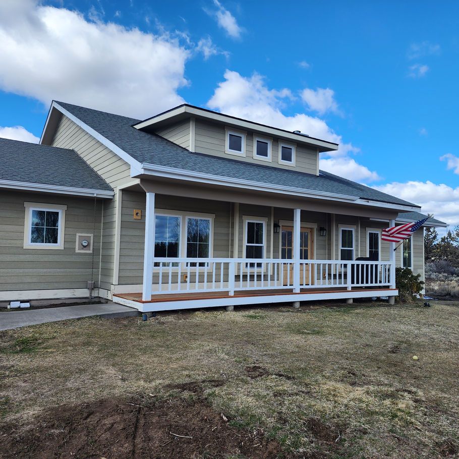 A single-story, light green house with a porch, white railings, and an American flag on a blue sky day.
