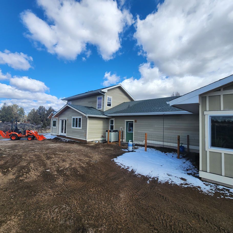 A two-story house with a green roof and tan siding sits on a dirt lot with patches of snow. Construction equipment is visible in the background under a blue sky.