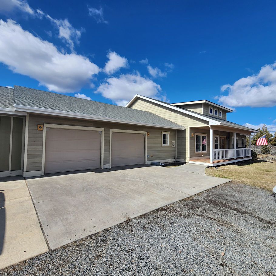A one-story house with a two-car garage, light-colored siding, and a covered porch under a blue sky with scattered clouds.