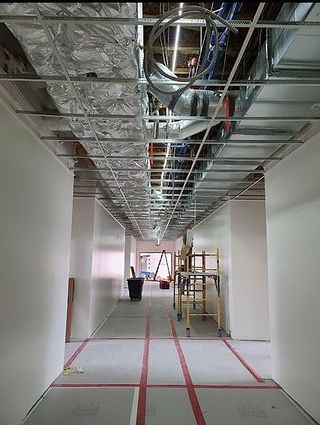 Interior hallway under construction with exposed ceiling beams and ductwork. Red tape marks floor, and there is construction equipment in the distance.