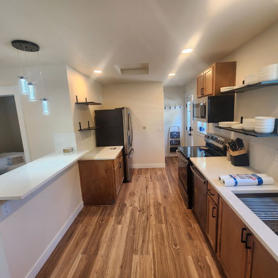 A narrow kitchen with wooden floors, wood cabinets, and white countertops. A stainless steel refrigerator stands between the cabinets.