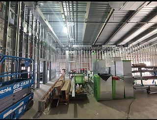 Interior of a construction site, featuring metal framing, ductwork, and construction materials. A scissor lift sits to the left.