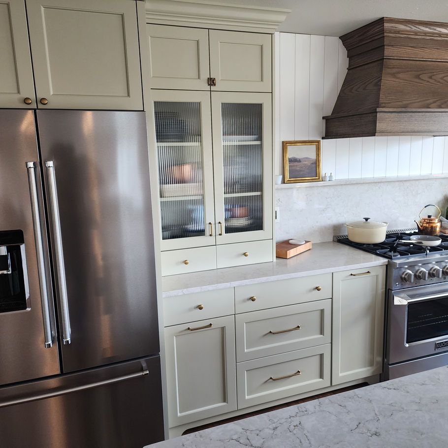 Kitchen with light green cabinets, stainless steel refrigerator, and wooden range hood. A built-in cabinet has glass doors.